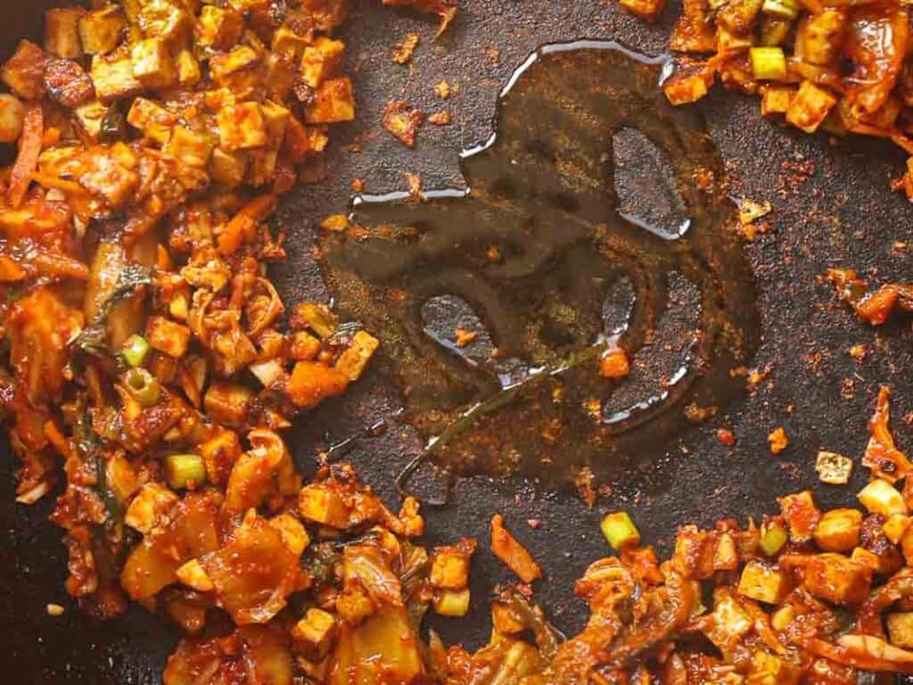 Close-up of a skillet with diced vegetables and tofu, sautéed in oil with spices. The mix is arranged in a ring around a bare center, revealing the pan’s surface.